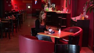Eine junge Frau sitzt allein in einer Bar und schaut wütend auf ihr Handy.