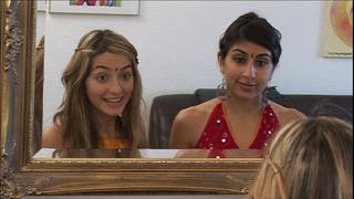 Zwei junge Frauen stehen vor einem Spiegel und sprechen miteinander, beide tragen einen Bindi.