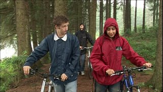 Vier junge Erwachsene schieben ihre Fahrräder durch einen Wald, es regnet.