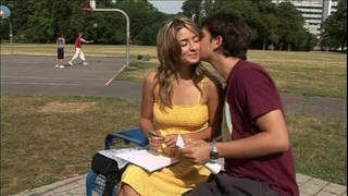 Ein junges Pärchen sitzt neben einem Basketballplatz, der junge Mann küsst die junge Frau auf die Wange.