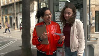 Zwei junge Frauen stehen vor einem Kiosk und schauen sich einen Lottoschein an.