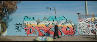 Zwei Menschen laufen im heutigen Berlin vor der Mauer, die mit Graffiti bespüht ist. Die Teilung Deutschlands hat ihr Leben geprägt.