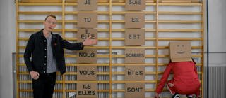 Ein junger Mann vor einer Sprossenwand, an ihr hängen Schilder. Auf den Schildern stehen französische Pronomen und die Konjugationen von "Être". Daneben sitzt eine Person mit Karton auf dem Kopf.