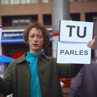 Ein junger Mann steht auf der Straße und hält Schilder hoch, darauf steht "Tu parles français"