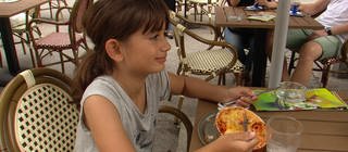 Ein Mädchen sitzt an einem Tisch im Biergarten und isst Lasagne. 