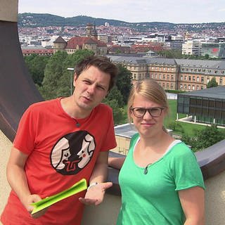 Ein Mann und eine Frau stehen vor einem runden Loch in einer Mauer, im Hintergrund eine Stadt.