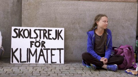 Greta Thunberg sitzt an einer Wand, neben ihr ein Plakat zu Klimastreik.