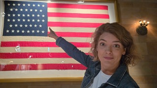 Host Lena Ganschow zeigt auf die amerikanische Flagge.